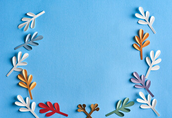 Frame of Fourteen leaves cut out of paper lie on a blue background with space for text in the center. The leaves are white, blue, green, red, brown and purple.  Flat lay, top view