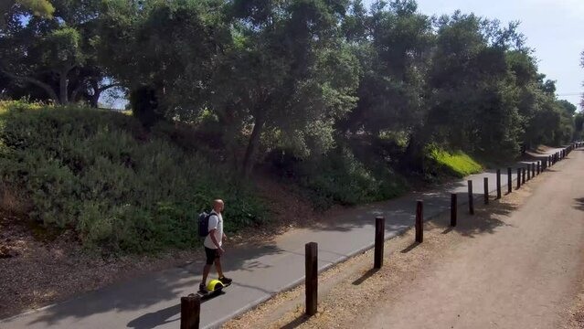 aerial footage of an African American man riding a Onewheel electric skateboard on a bike trail surrounded by lush green trees and grass and dry brush with blue sky at Duarte California USA