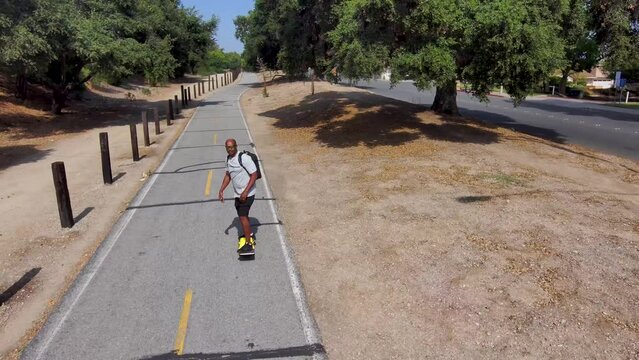 aerial footage of an African American man riding a Onewheel electric skateboard on a bike trail surrounded by lush green trees and grass and dry brush with blue sky at Duarte California USA
