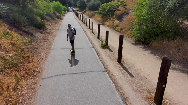 aerial footage of an African American man riding a Onewheel electric skateboard on a bike trail surrounded by lush green trees and grass and dry brush with blue sky at Duarte California USA