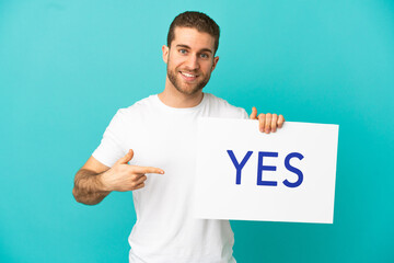 Handsome blonde man over isolated blue background holding a placard with text YES and  pointing it