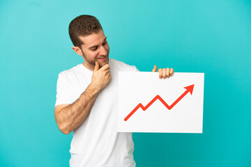 Handsome blonde man over isolated blue background holding a sign with a growing statistics arrow symbol and thinking