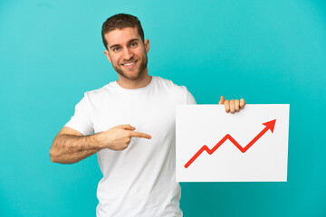 Handsome blonde man over isolated blue background holding a sign with a growing statistics arrow symbol and  pointing it