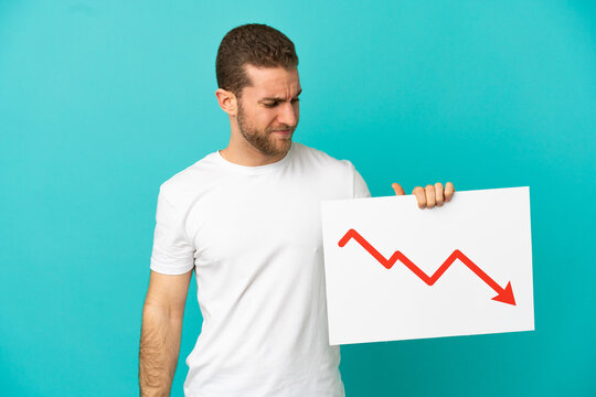Handsome blonde man over isolated blue background holding a sign with a decreasing statistics arrow symbol with sad expression