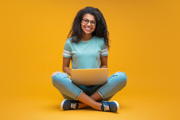 Studio portrait of smiling young beautiful african american girl in trendy spectacles, sitting with laptop and looking directly to the camera, isolated over bright orange yellow background