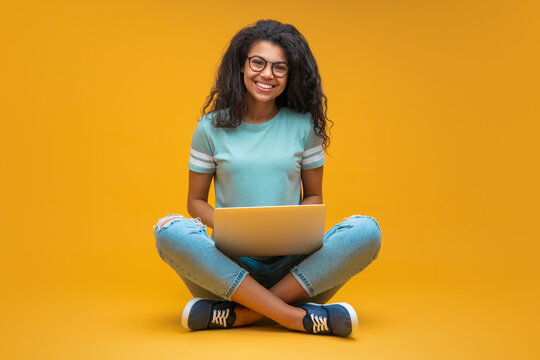 Studio portrait of smiling young beautiful african american girl in trendy spectacles, sitting with laptop and looking directly to the camera, isolated over bright orange yellow background