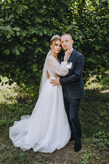 A young groom in a blue plaid suit and a beautiful bride in a white lace dress with a diadem, a bouquet in her hand are gently hugging in a park in nature. Wedding photo of smiling newlyweds.