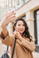 Glamour attractive asian kazakh female wearing beige coat, taking selfie, checking haircut, look at smartphone camera, walking on street