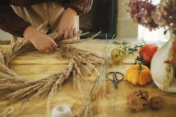 Making stylish autumn wreath on rustic table. Woman hands arranging dried grass in wreath on wooden table with scissors, thread, pumpkin. Fall decor and arrangement in farmhouse.