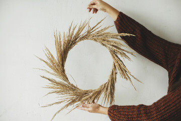 Woman hands holding rustic wreath on white wall background. Stylish boho autumn wreath with dried grass. Fall decor and arrangement in farmhouse