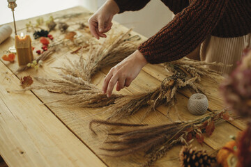Making autumn wreath on rustic table. Stylish woman arranging dried grass in wreath on wooden table...