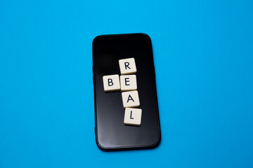 Be real text with plastic letters on mobile phone and blue paper. Popular and trendy mobile app called Be Real is spreading around the globe. Be realistic yourself is a good way of living your life.