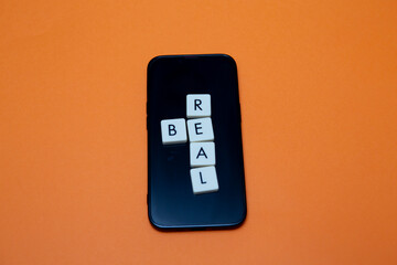 Be real text with plastic letters on orange and mobile phone. Popular and trendy mobile app called Be Real is spreading around the globe. Be realistic yourself is a good way of living your life.