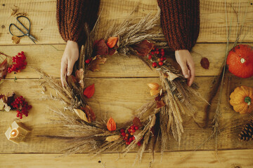 Autumn wreath flat lay on rustic table. Woman hands holding stylish wreath with berries, dry grass,...
