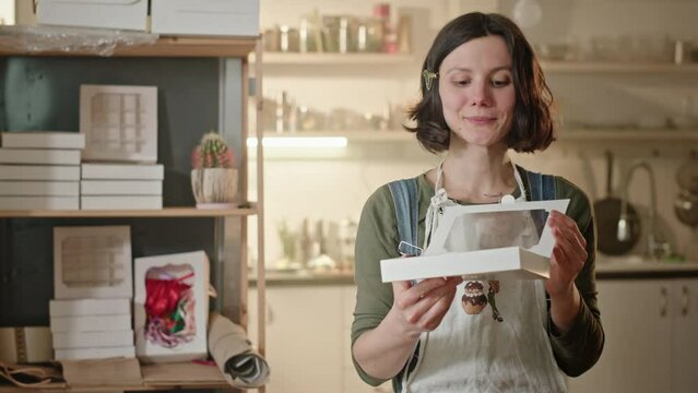 Young brunette housewife demonstrates box of cookies smiling. Young brunette housewife in apron holds white dessert box in hands. Woman opens box and demonstrates homemade cookies smiling happily in