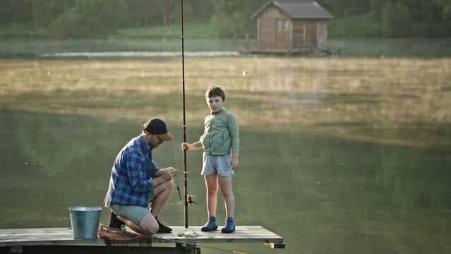 Fisherman squats on pier and shows schoolboy to hook bait. Fisherman squats on river wooden pier near bucket on sunny summer day. Father adjusts line and shows son to hook bait. Child looks at bait