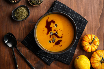One deep black bowl with an orange pumpkin cream soup with pumpkin seed oil, two black spoons on a...