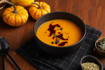 One deep black bowl with an orange pumpkin cream soup with pumpkin seed oil, two black spoons on a wooden surface
