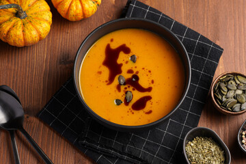 One deep black bowl with an orange pumpkin cream soup with pumpkin seed oil, two black spoons on a wooden surface