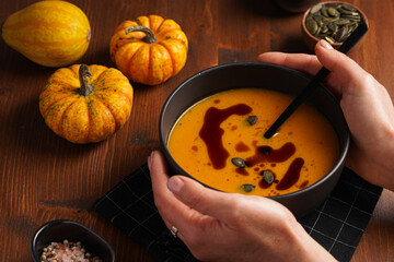 Female hands holding one deep black bowl with an orange pumpkin cream soup with pumpkin seed oil,...