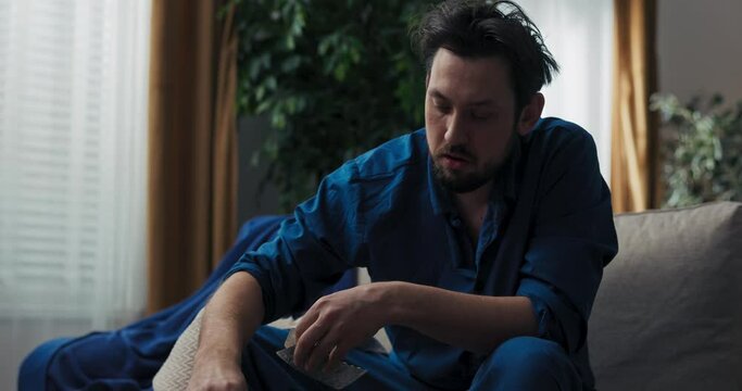 Close-up of man in blue pajamas sitting on bed and taking pills. He has fever. He holds the pills in hands and chooses the right one. The man press the pill on hand and drinks it with tea.