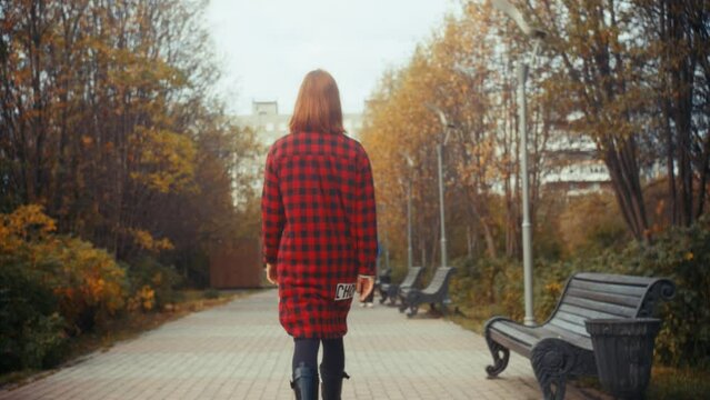Following with rotation a young slim redhead caucasian girl in a red and black plaid coat and black boots in a city park with benches, lanterns and trees with yellow leaves on an autumn cloudy day
