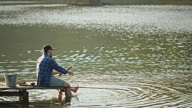 Fisherman sits on pier with rod in hand and spins reel. Fisherman sits on wooden pier with rod in hand near bucket and net fishing in sunny morning. Man spins reel and swings legs in water side view.