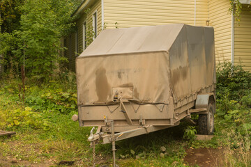 a modern trailer covered with an awning, stained with road dirt. the background of a country house and foliage.
