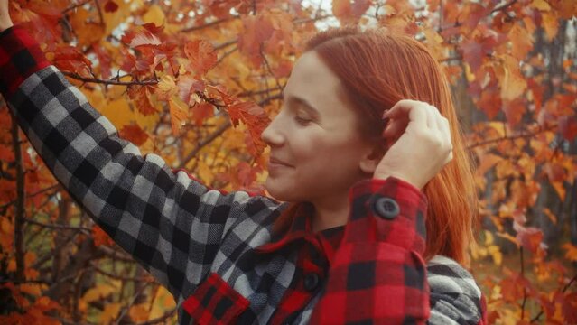 A beautiful young caucasian redhead girl in a red-black plaid coat fooling around and play with an orange leaf among the trees, puts it on her ear and head in a city park on an autumn cloudy day