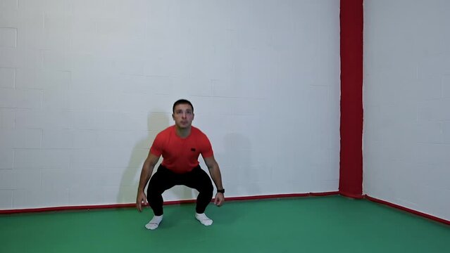 Nice slow motion footage with young caucasian male athlete trains by performing squat jump exercise. Background with white wall, copy space.