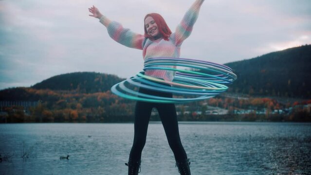 A young beautiful smiling redhead girl in black leggings and a multi-colored jacket spins hoops at waist near lake and hills in autumn evening. Exercise from rhythmic gymnastics and than pan on sky