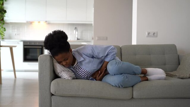 Exhausted African American woman lying on sofa grabbing belly experiencing pain and heaviness in stomach. Black female in casual clothes suffering from health problems due to eating junk fast food 