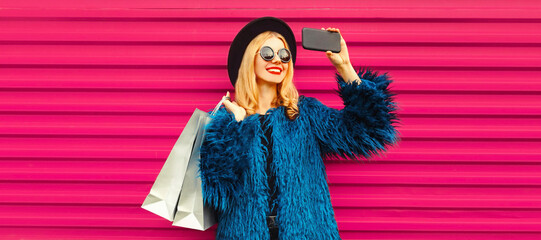 Portrait of happy smiling blonde woman taking selfie with smartphone with shopping bags wearing blue fur coat, black round hat on pink background