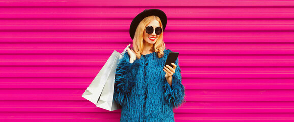 Portrait of stylish blonde woman with smartphone and shopping bags wearing blue fur coat, black round hat on colorful pink background