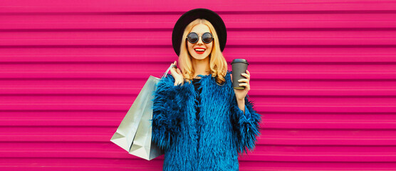 Portrait of beautiful stylish smiling woman with shopping bags wearing blue fur coat, black round hat and sunglasses posing on pink background