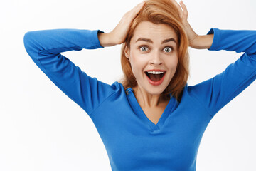 Close up portrait of woman looking surprise, holds hands on head and staring amazed at camera, impressed by smth awesome, stands over white background