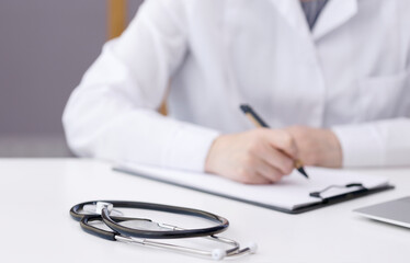 A woman in a white medical coat sits at a white table and writes on paper. The doctor in the hospital office works with papers and patients. There is a stethoscope on the table. High quality photo