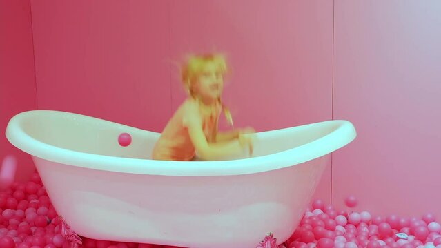 Happy Little pretty blond caucasian girl stands by the bathroom and throws pink balls.