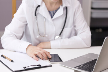 The doctor at the white table looks at the information on the phone. A female doctor works in a hospital office. A doctor in a white coat sits in front of a laptop and looks for information on the