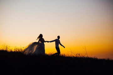 silhouette of a cheerful couple, the bride and groom in a wedding dress, laughing and holding hands, run across the field