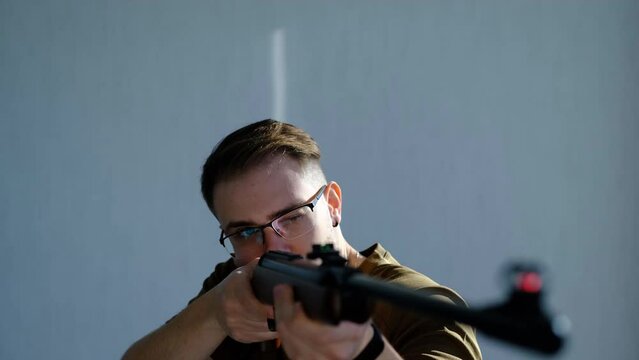 a young man in glasses looks in aim of air gun and look for prey on a gray background. Hunting with air gun. Forward view