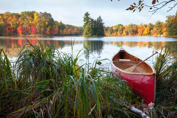 Red wooden canoe on shore of lake with trees in autumn color and a small island - Powered by Adobe