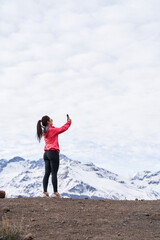 young woman standing in red jacket, taking a picture with her phone in the middle of the Andes Mountains of Chile