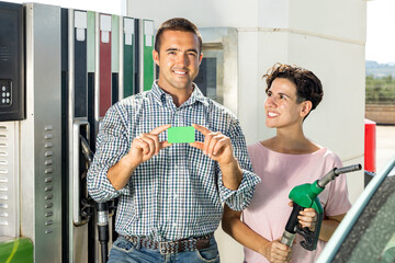 Smiling man and woman demonstrating gas credit card while filling car tank in petrol filling station