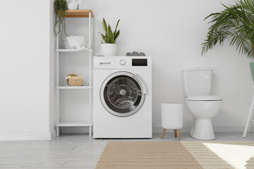 Modern washing machine, shelf unit and toilet bowl near white wall in bathroom interior