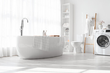 Stylish interior of light bathroom with bathtub, shelf unit and washing machine