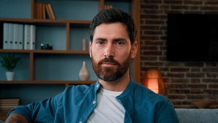 Head shot handsome bearded caucasian guy confident serious 30s male portrait wears casual blue shirt attractive caucasian man homeowner client sit on sofa at home looking at camera with serene emotion
