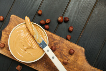 Board with bowl of tasty hazelnut butter on dark wooden background, closeup