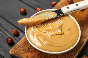 Board with bowl of tasty hazelnut butter on dark wooden background, closeup