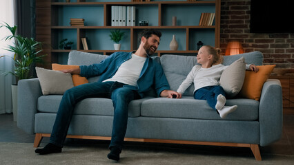 Happy caucasian young family daddy and child daughter jumping on soft couch for daytime break relaxation laugh enjoy relax rest on cozy couch testing comfort of sofa in furniture store room interior
